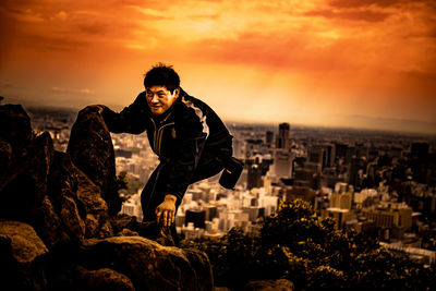 Man standing on rock by cityscape against sky during sunset