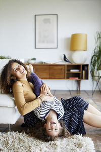 Happy mother holding daughter upside down while sitting on floor by sofa at home
