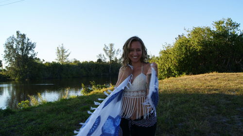 Portrait of smiling young woman holding scarf by lake on field