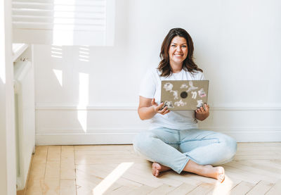 Smiling brunette woman in casual clothes plus size body positive using laptop sitting in bright room