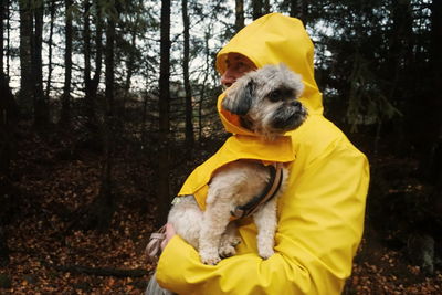 Man wearing yellow raincoat carrying dog in forest