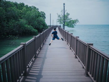 Boy jumping on walkway over sea against sky