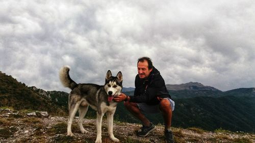 Smiling mature man pampering dog on mountain peak against cloudy sky