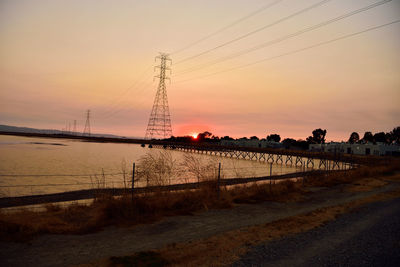 Silhouette electricity pylon by sea against sky during sunset