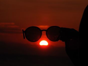 Silhouette hand holding sunglasses against sky during sunset