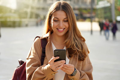 Young cheerful stylish woman using cell phone and texting message on city street. 