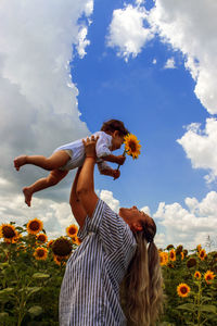 Mother holding boy with sunflower at field against sky