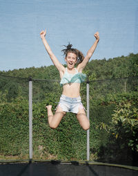 Vertical photo of a smiling young brunette teenager jumping on a trampoline with net around on the green yard outdoors. the girl wears short jeans and a top and looks fun, active