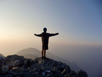 Rear view of man with arms outstretched standing on rock against sky