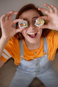 From above delighted young female keeping tasty sushi near eyes while having lunch at home