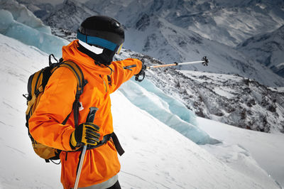 A professional skier athlete in a red jacket, helmet and mask stands on a slope against the backdrop