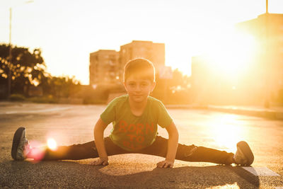 Portrait of boy stretching legs in city against sky during sunset