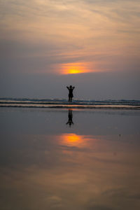 Mid distance view of woman standing on shore at beach against sky during sunset