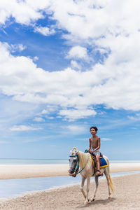 Young boy riding horse on the beach with blue sky and ocean in background.