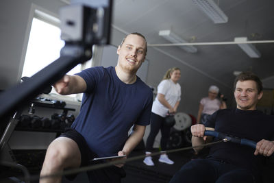 Smiling male fitness instructor looking at rowing machine by man exercising in health club