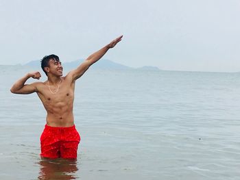 Smiling shirtless man gesturing while standing in sea against sky