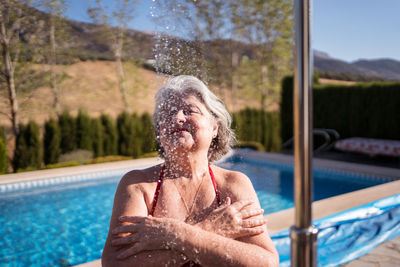 Cheerful senior female in bikini enjoying splashes from shower near pool with transparent clear water