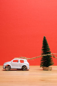 Close-up of small christmas tree tied with toy car on wooden table against red background