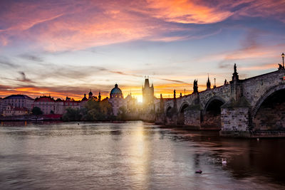 Arch bridge over river amidst buildings against sky during sunset