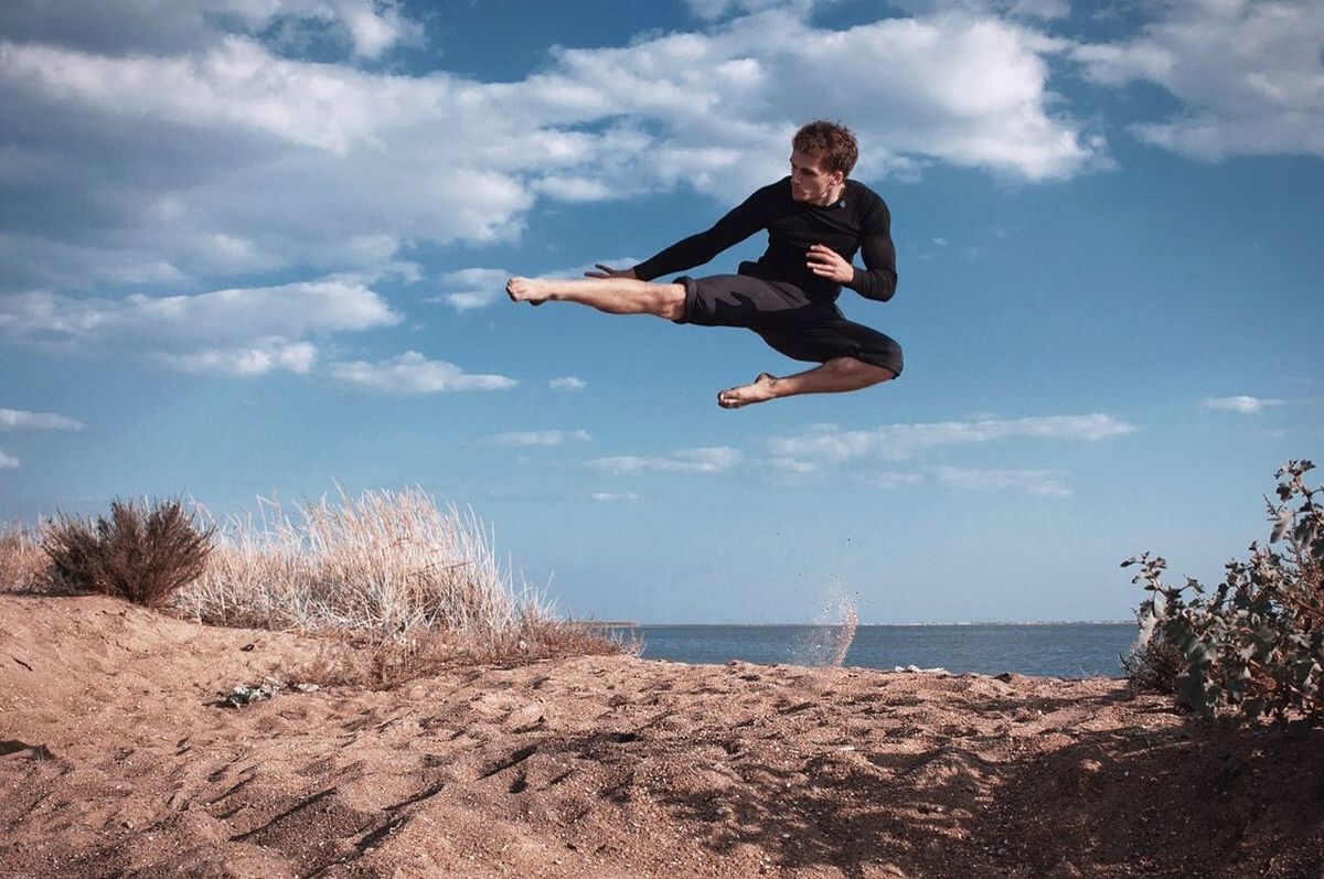 full length, lifestyles, leisure activity, person, sky, young adult, casual clothing, mid-air, jumping, fun, enjoyment, sea, young men, beach, water, vacations, cloud - sky, arms outstretched