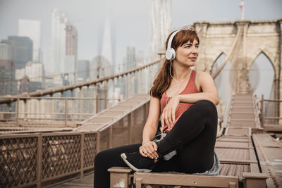 Woman listening to music while sitting on bridge against sky