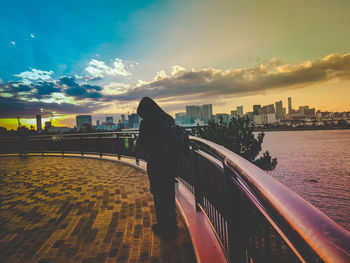 Man standing by railing in city against sky during sunset