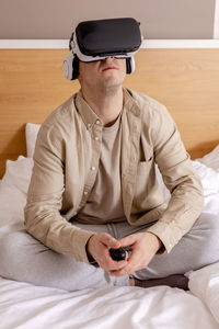 Young caucasian man sitting on bed at home with vr headset and playing interactive video game
