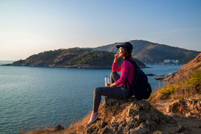 Rear view of woman sitting on rock by sea against sky