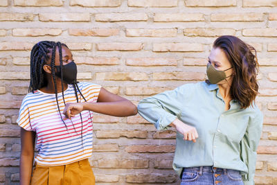 Smiling friends wearing mask doing elbow bump while standing against wall outdoors