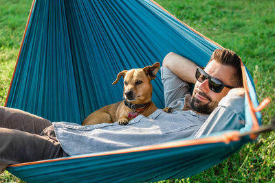 Young handsome european man in sunglasses is resting in hammock with his cute little dog.