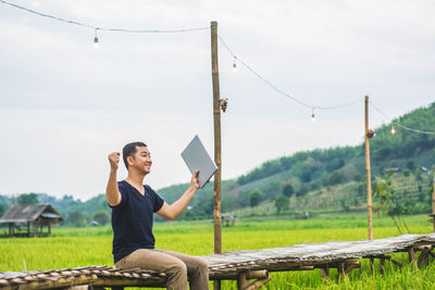 Man with arms outstretched holding laptop while sitting on boardwalk at rice paddy