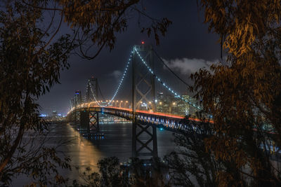 Illuminated bay bridge over river against sky at night