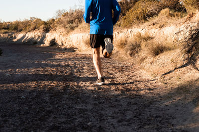 Back view of unrecognizable active male jogger running uphill in sandy semi desert terrain during workout in sunny morning