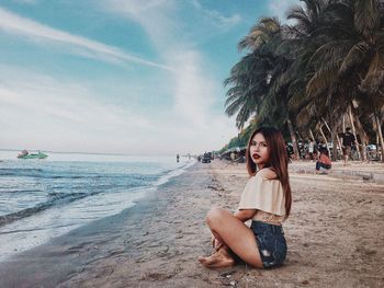 Young woman sitting on palm tree at beach against sky