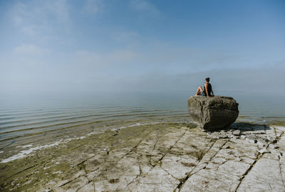 Woman looking at sea while sitting on rock against blue sky