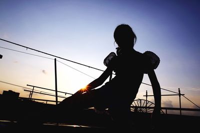 Silhouette woman sitting on railing against sky during sunset