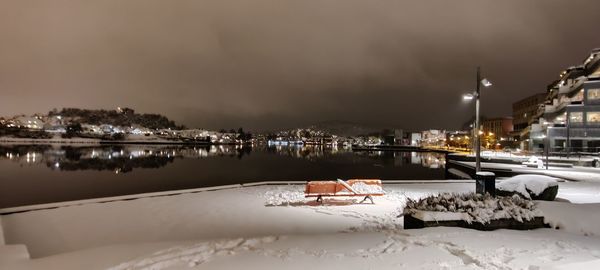 Illuminated buildings by frozen river against sky at night