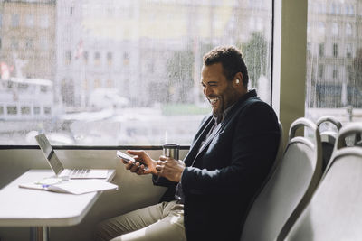 Cheerful male entrepreneur with mobile phone and insulated drink container sitting at table in ferry