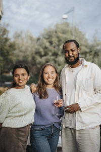 Portrait of smiling male and female friends standing together while holding wineglass during dinner party at cafe