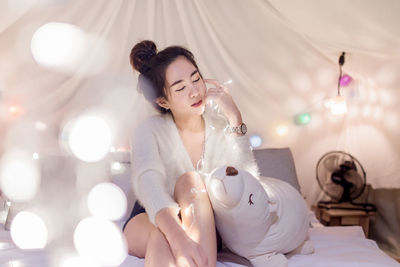 Young woman sitting with toy by illuminated lighting equipment on bed