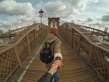 Cropped hand of man holding woman while walking on brooklyn bridge against sky