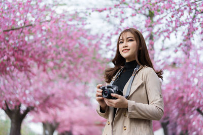 Portrait of beautiful young woman photographing with pink camera standing against trees