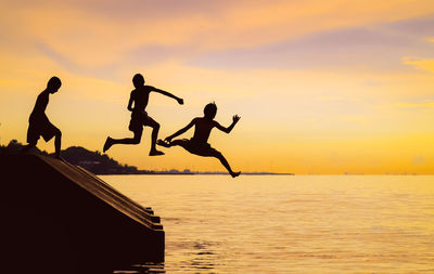 Silhouette people jumping over sea against sky during sunset