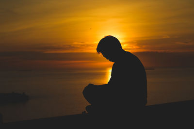 Silhouette man sitting on beach against orange sky