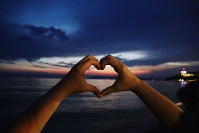 Hand holding heart shape against sky during sunset