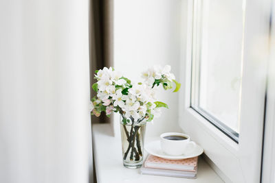 White cup of coffee on a white plate, a stack of books and a vase of flowers on the windowsill 