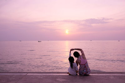 Rear view of mother with daughter sitting on footpath by sea against sky during sunset