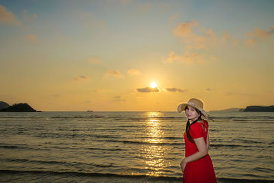 Portrait of young woman standing at beach against sky during sunset