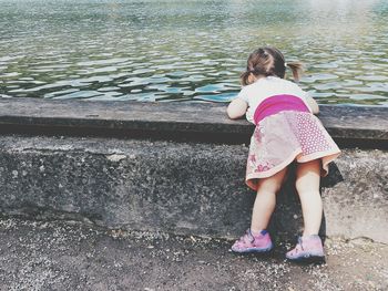 Rear view of girl leaning on retaining wall by pond