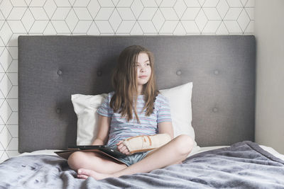 Young girl sitting on bed at home with a broken arm and device